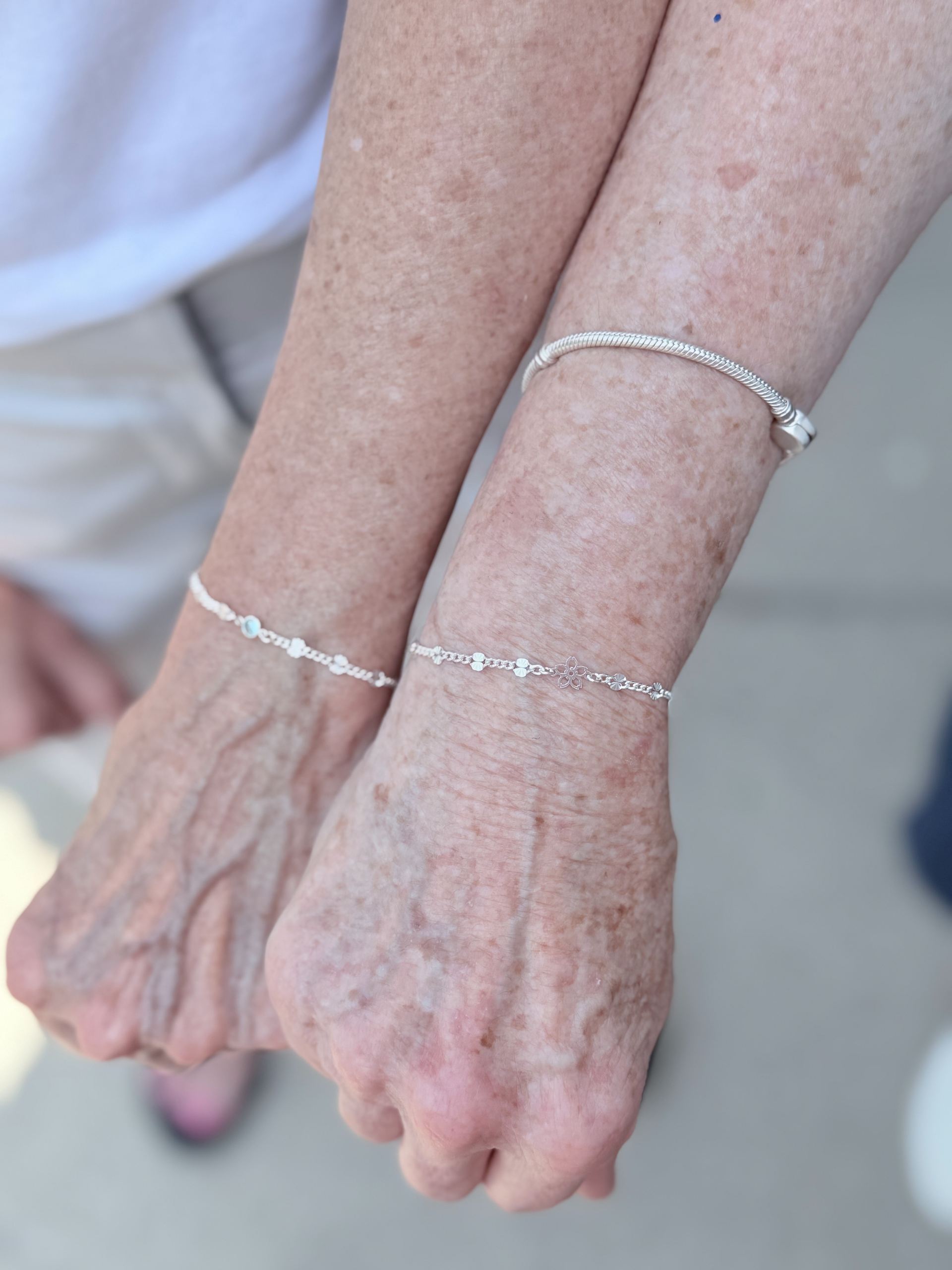Two wrists wearing matching silver bracelets with small flower and disc charms