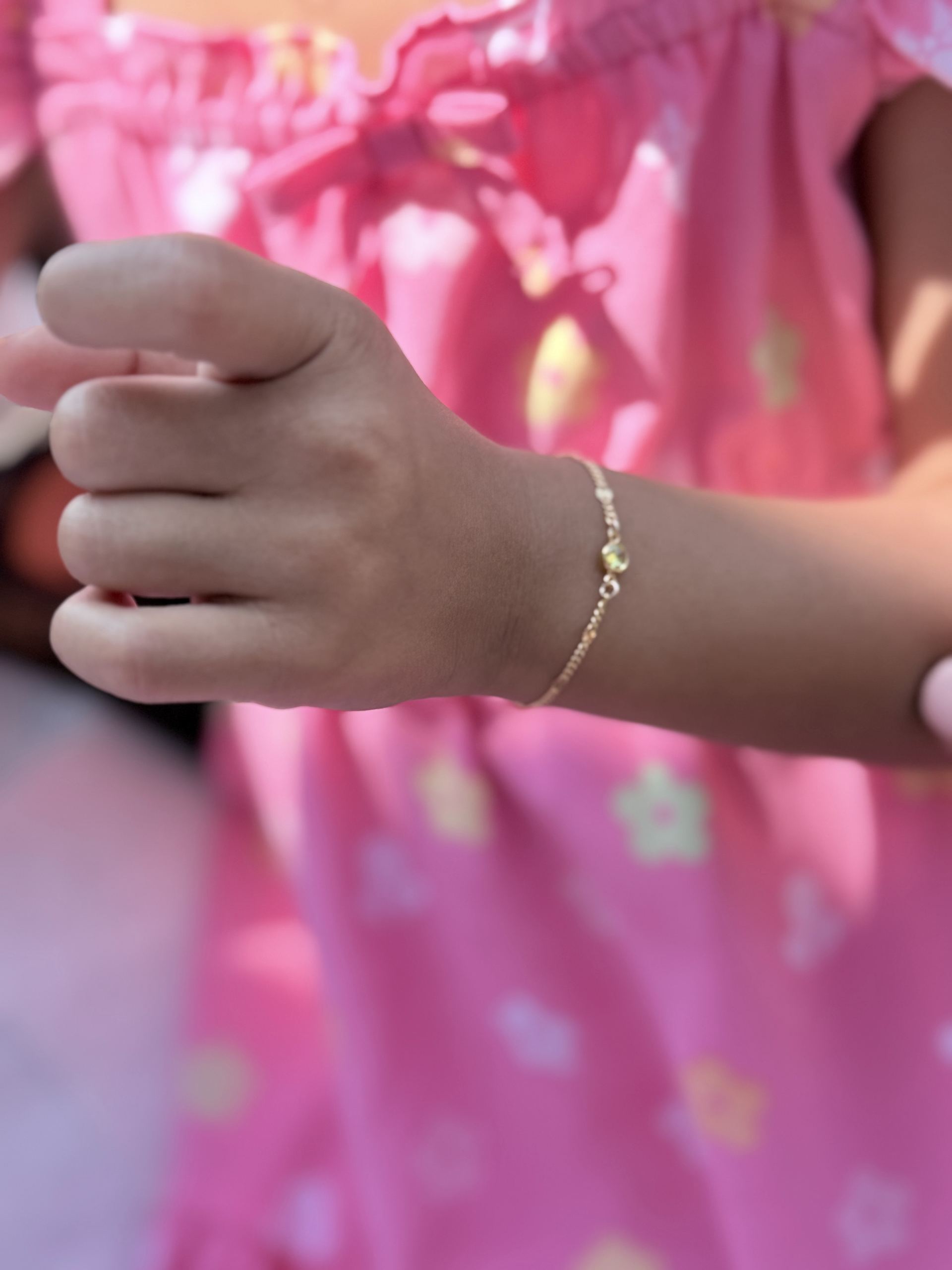 Child's wrist wearing a delicate gold chain bracelet with a single round gemstone charm