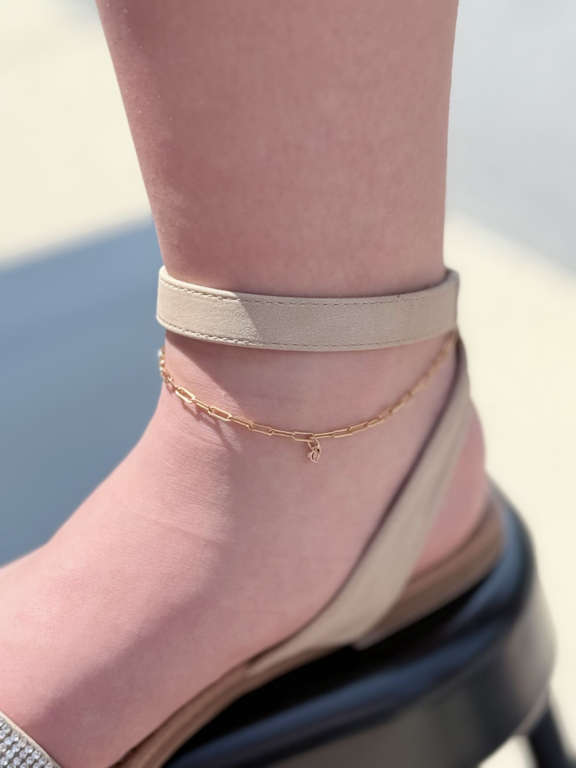 Gold paperclip-link chain anklet layered over a beige ankle-strap sandal