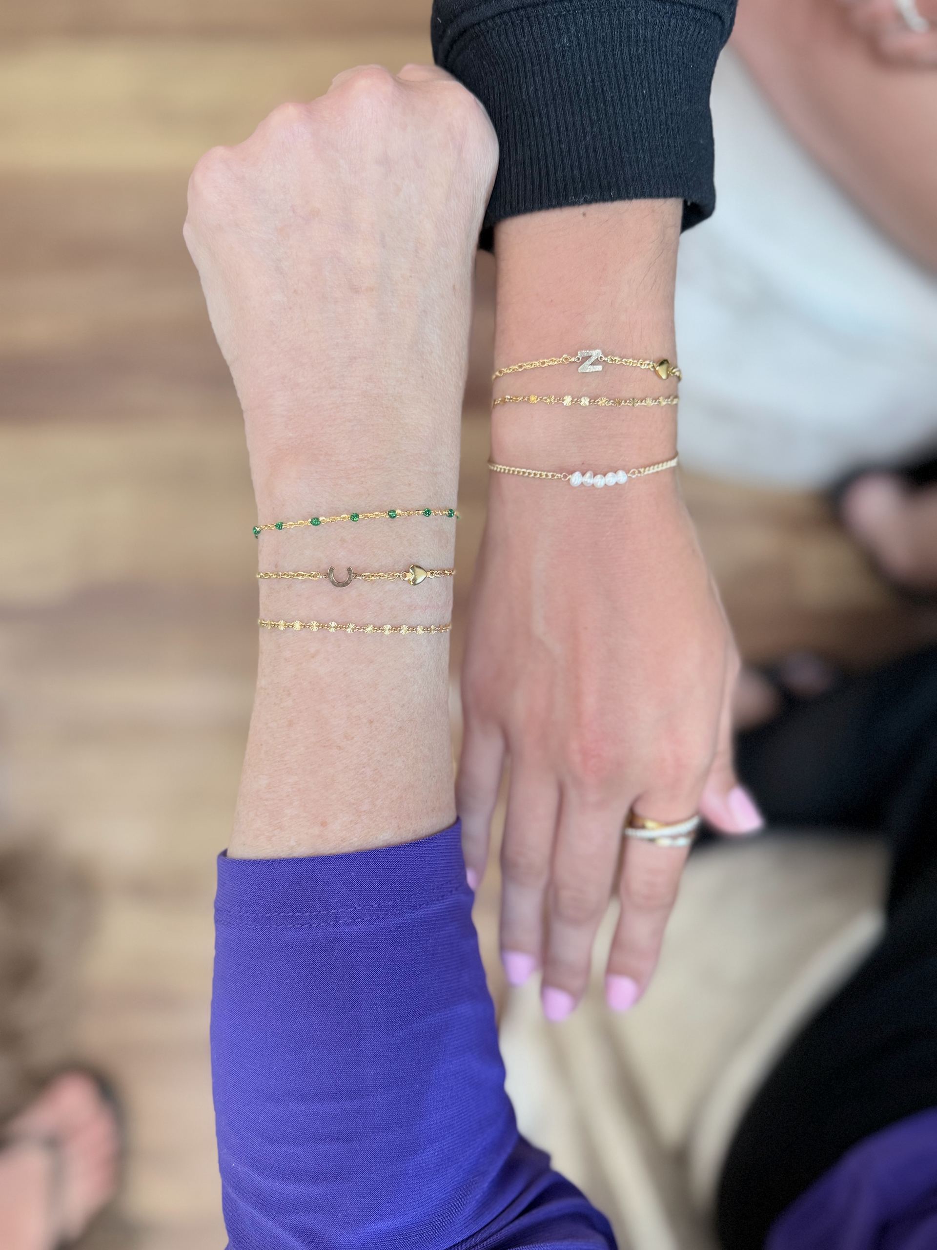 Two wrists layered with gold bracelets, one with a green-beaded chain and a horseshoe-and-heart charm, the other with a diamond letter Z charm and a seed-pearl bar bracelet