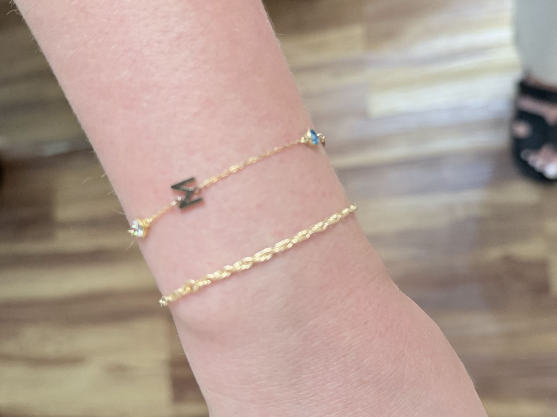 Gold bracelet with a blue oval gemstone bead and a small letter M charm, layered with a thinner gold chain