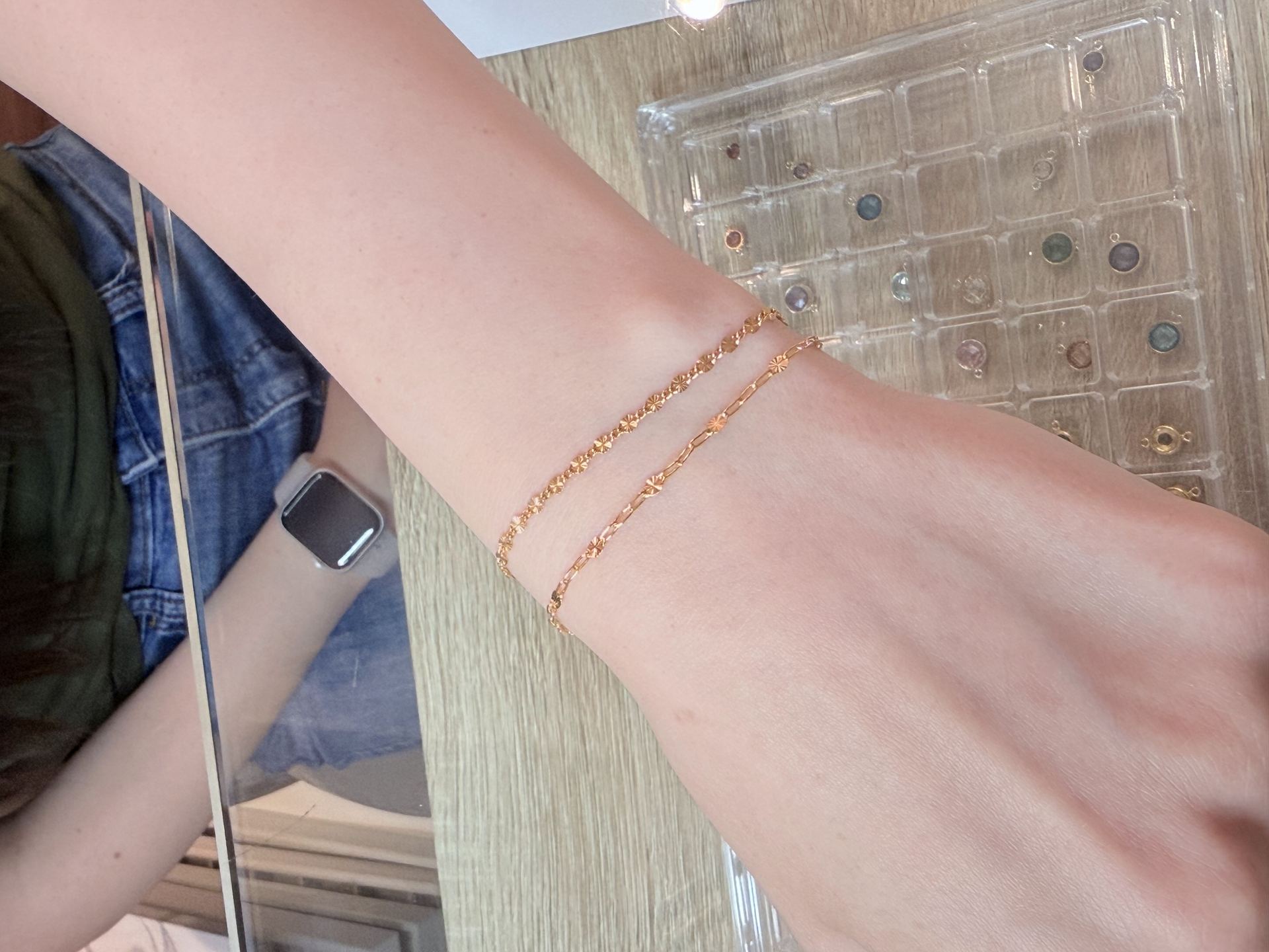Wrist wearing two rose-gold-tone bracelets, a textured chain and a paperclip-link chain, resting on a wooden surface