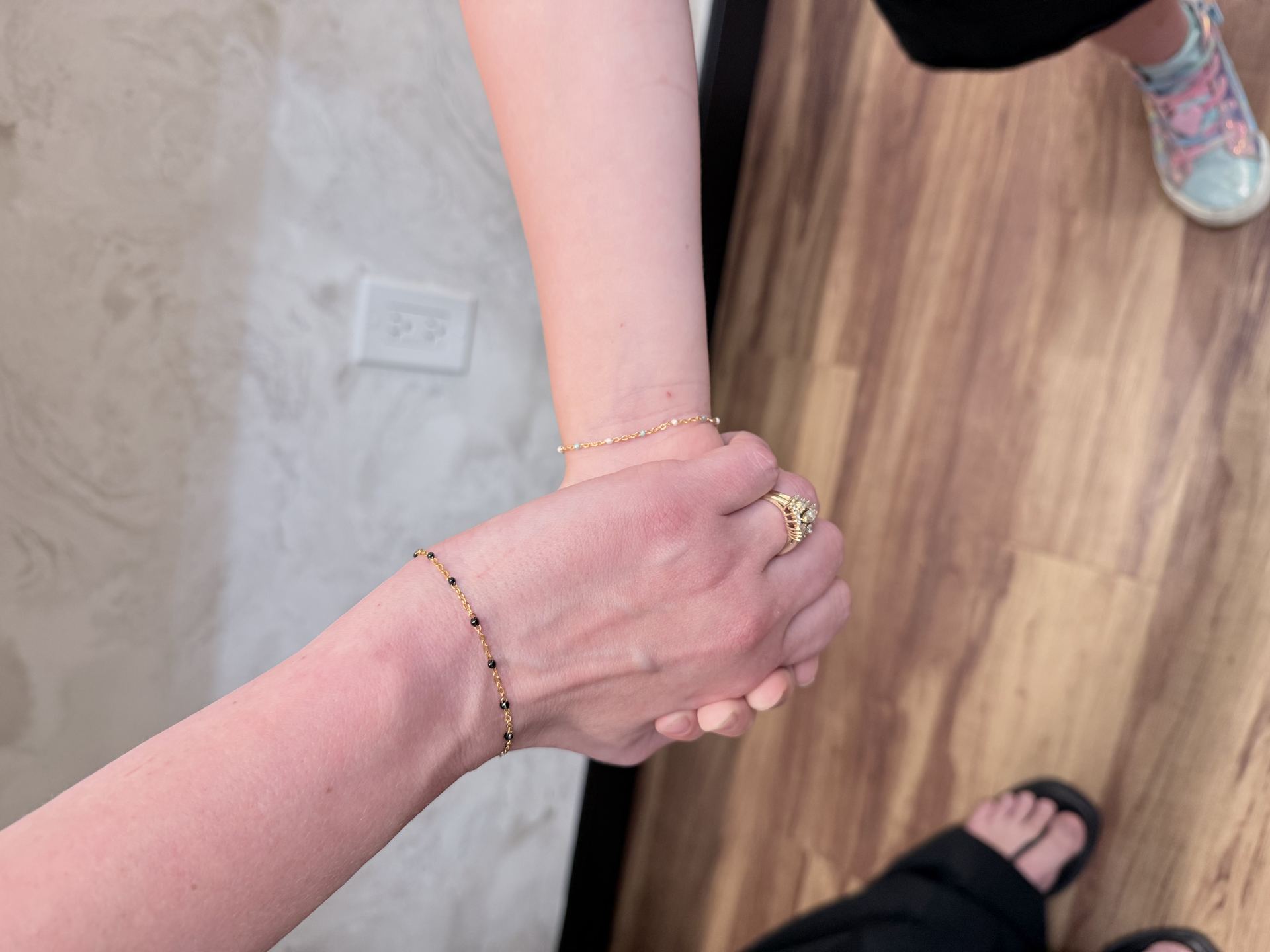 Two friends' wrists clasped together, each wearing a delicate gold chain bracelet with black beads