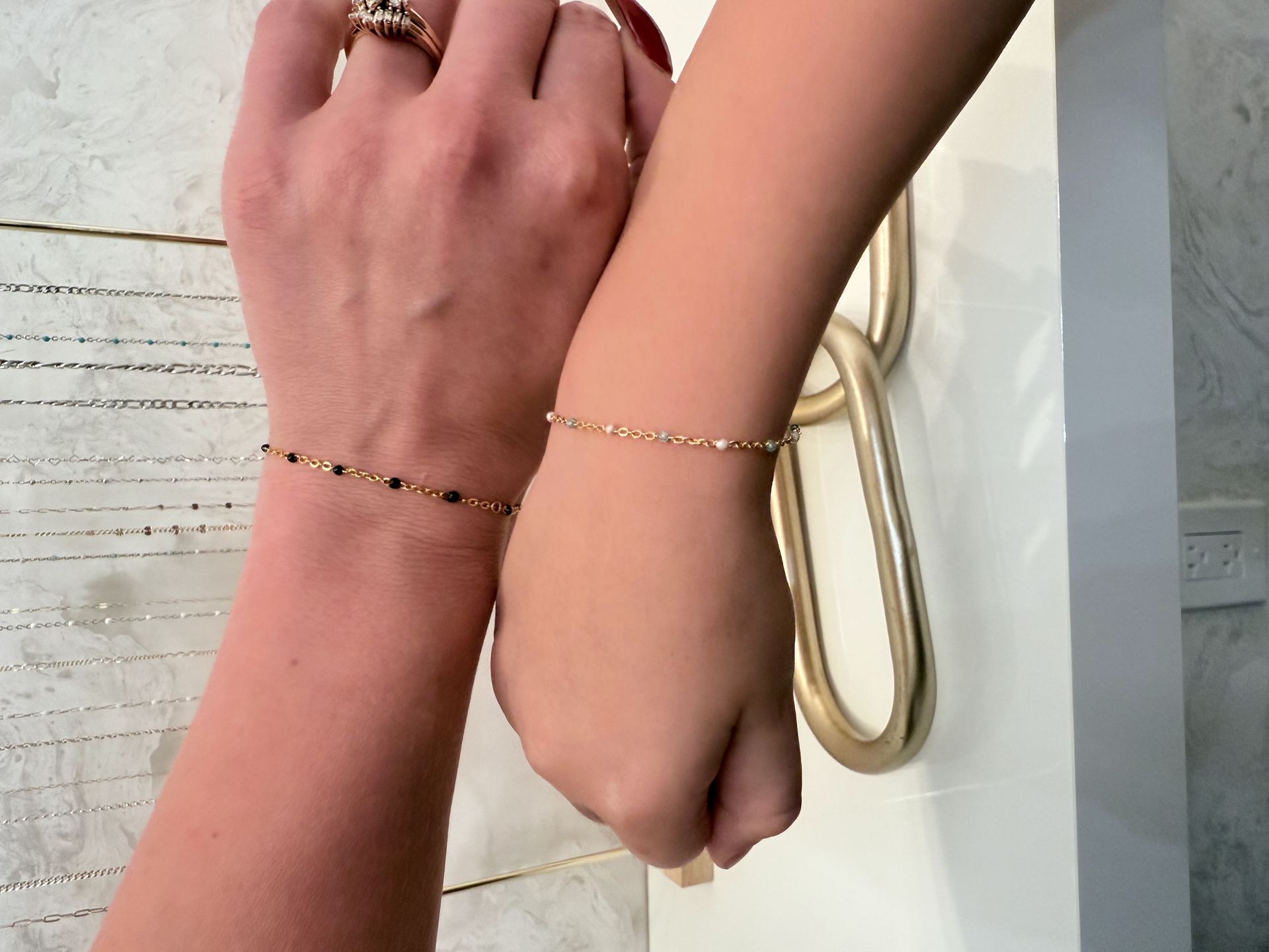 Two wrists pressed together wearing matching gold bead-and-chain bracelets, one with black beads and one with white beads