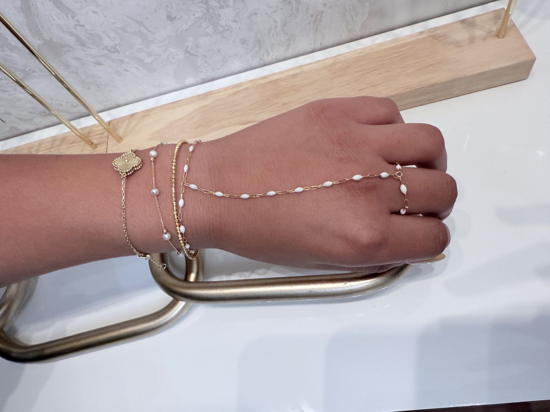 Hand and wrist layered with gold bracelets, a clover charm, a pearl chain, and a beaded chain, with rows of chain visible on a display board behind