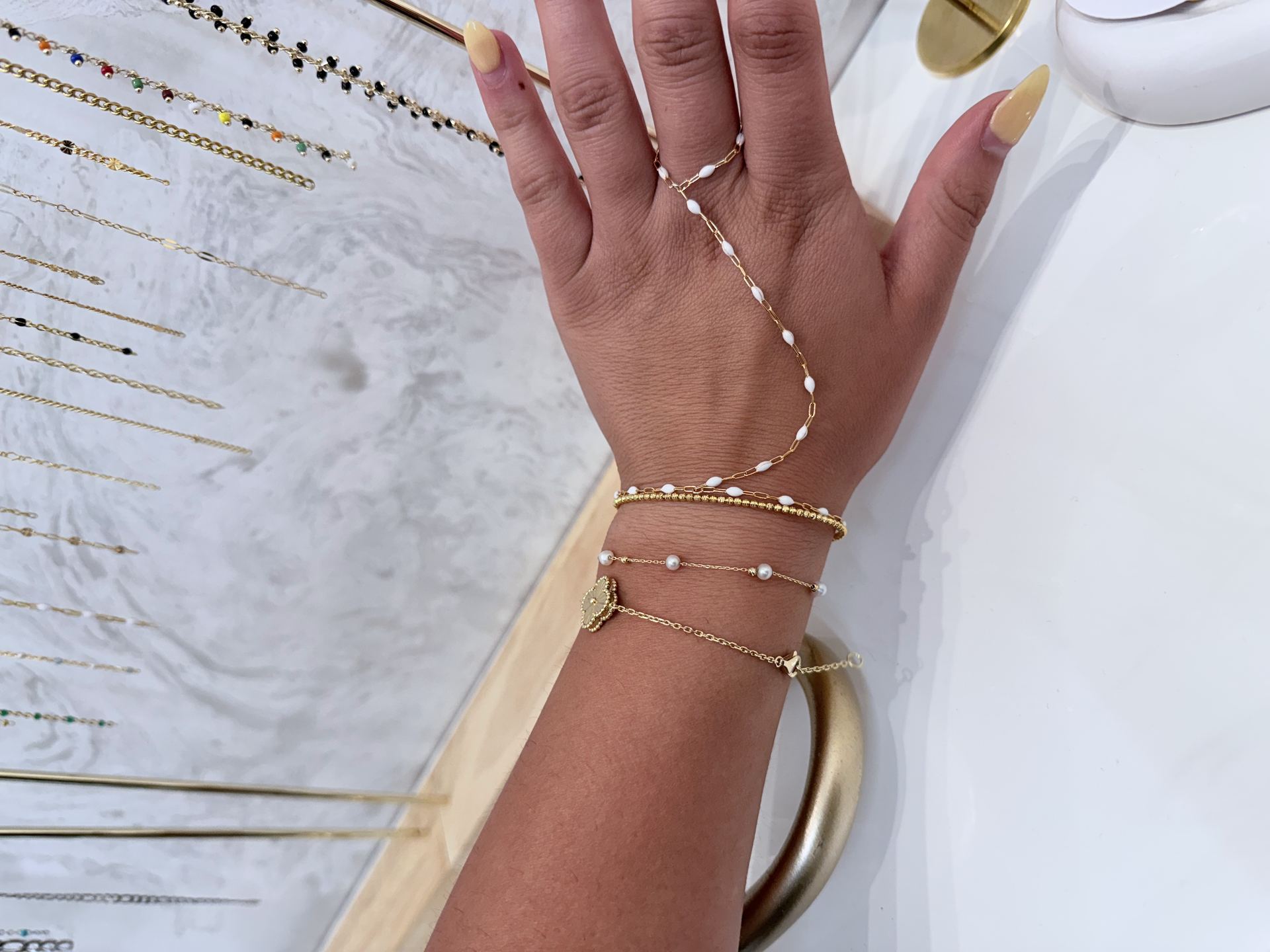 Hand wearing a white beaded chain draped from a ring across the fingers to a stack of gold bracelets including a clover charm and pearl chain