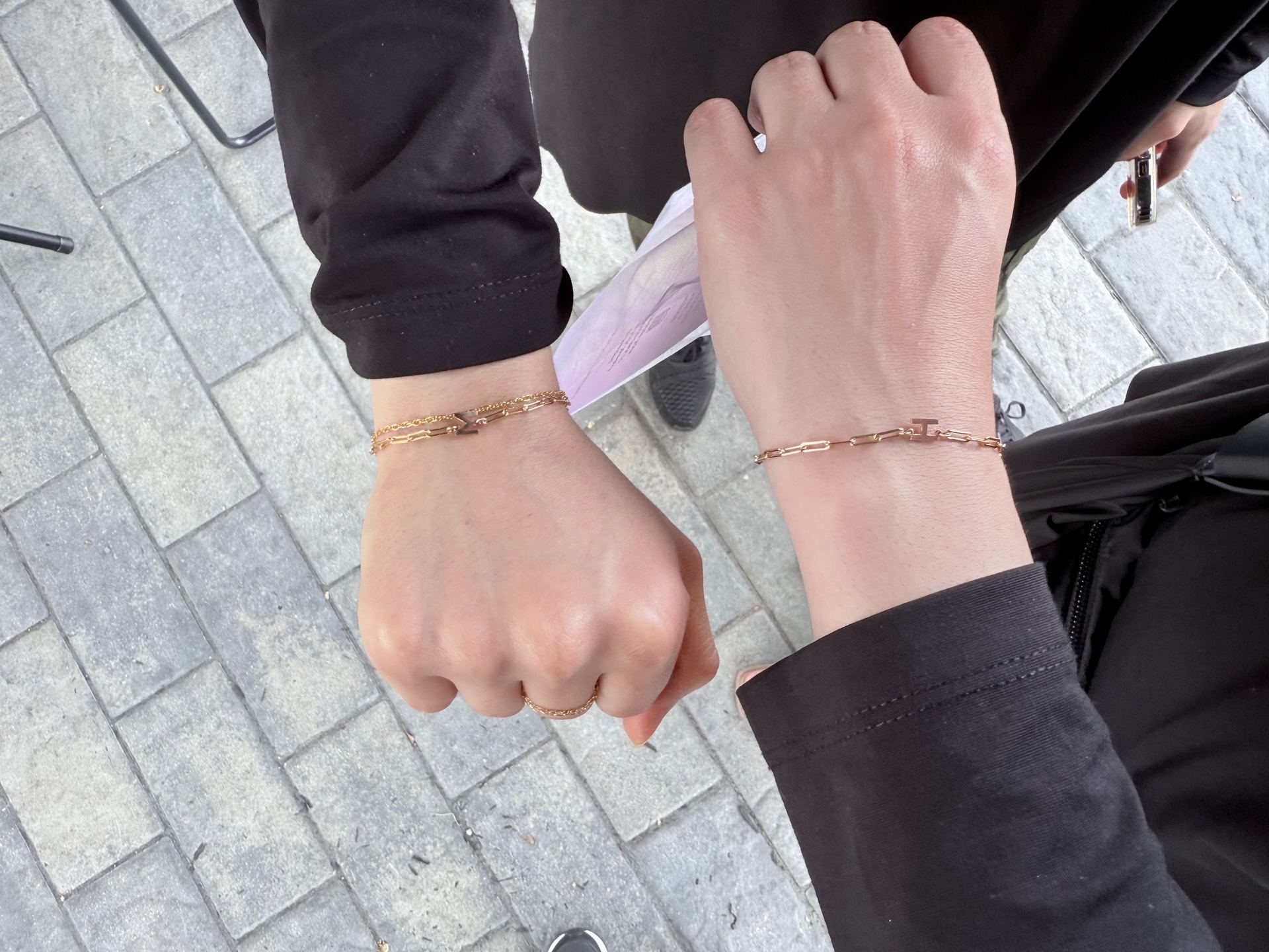 Two wrists fist-bumping, each wearing a gold paperclip-link bracelet with a letter charm, one reading M and the other T