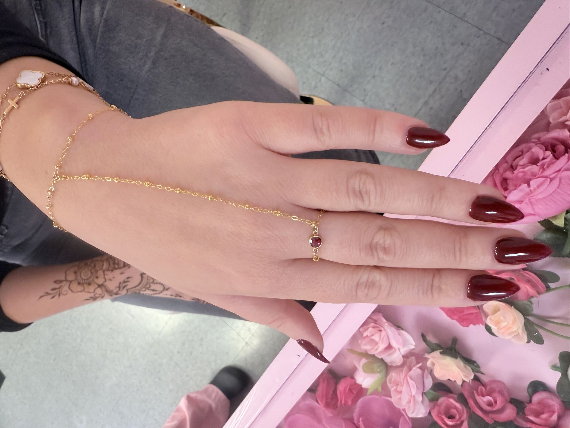 Hand wearing a gold chain hand harness with a round pink gemstone ring, connected to layered bracelets with a cross charm and a clover charm