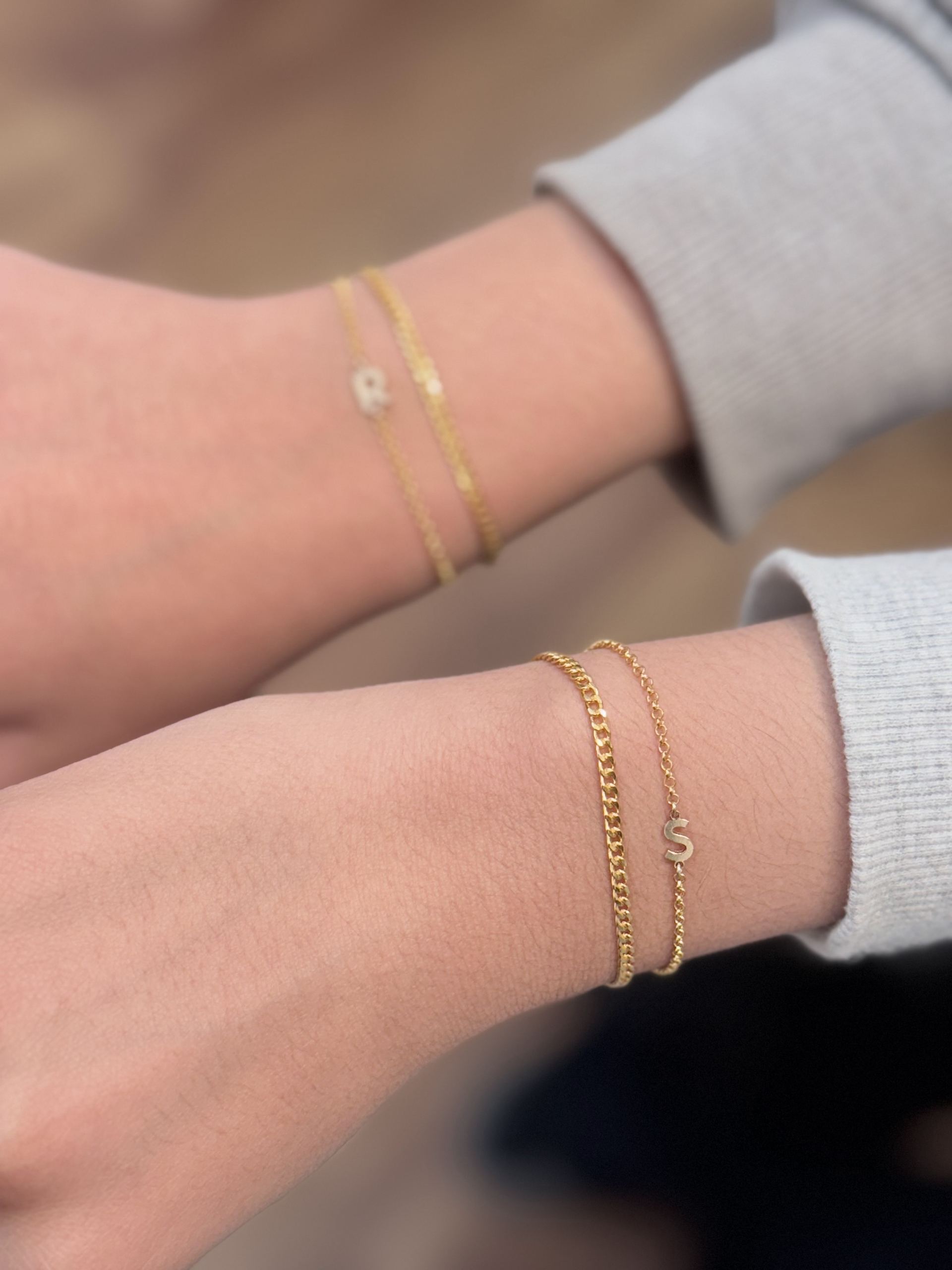 Two wrists wearing matching gold curb-chain bracelets with letter charms, one reading R and the other S