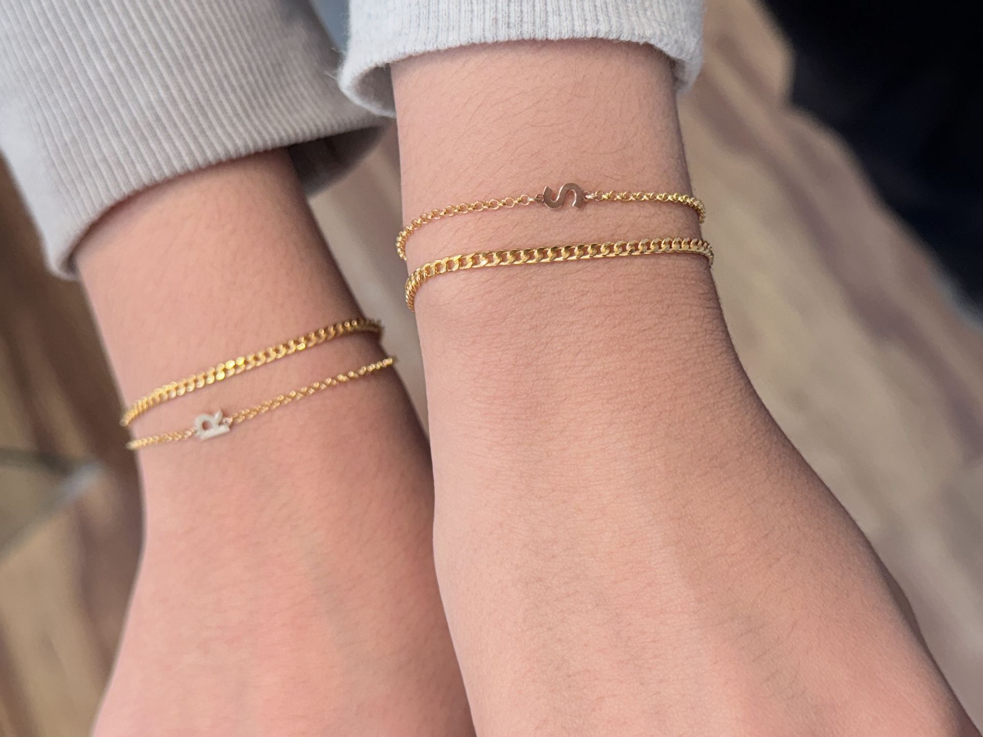Close-up of two wrists wearing matching gold curb-chain bracelets with letter charms reading R and S